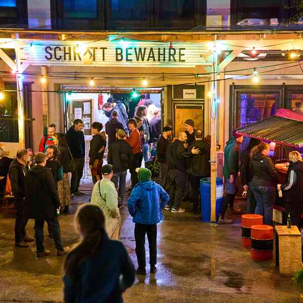 Der Eingang von Schrott bewahre in Abendstimmung. Eine Lichterkette leuchtet, Menschen stehen herum und unterhalten sich auf der Einfahrt. Eine Rampe führt in den Keller durch das geöffnete Garagentor.