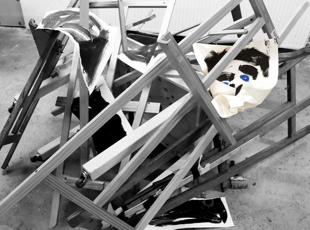 Ein Haufen zerschlagener und ineinander geworfener Bildstaffeleien liegt auf dem Boden. Ein Bild liegt in Fetzen verteilt über dem Haufen. Die Aufnahme in schwarz-weiß Tönen gehalten.