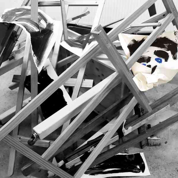 Ein Haufen zerschlagener und ineinander geworfener Bildstaffeleien liegt auf dem Boden. Ein Bild liegt in Fetzen verteilt über dem Haufen. Die Aufnahme in schwarz-weiß Tönen gehalten.