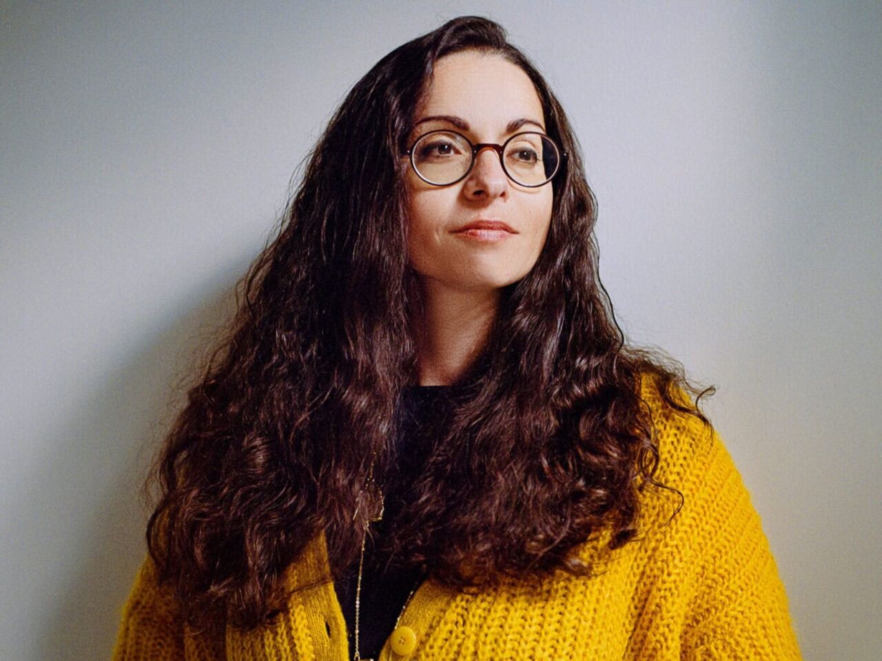 Porträt von Marina Chernivsky in gelber Strickjacke mit offenen braunen Locken und Brille.